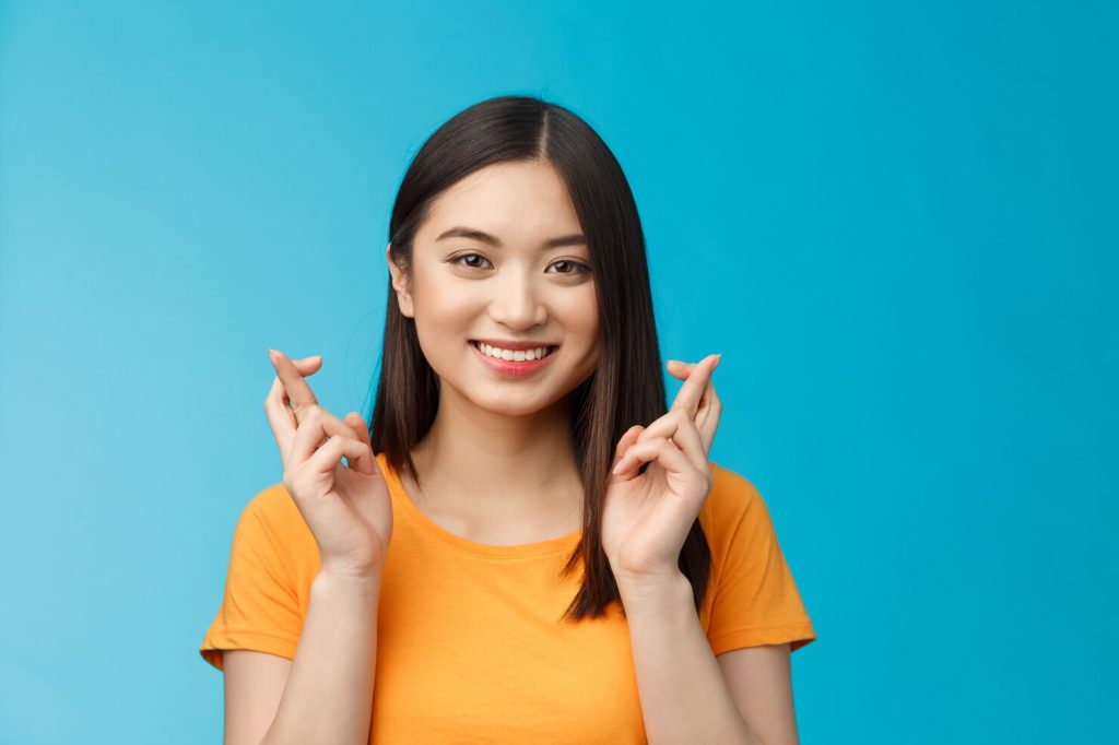 close up hopeful cute asian girl with short dark hair cross fingers good luck praying smiling broadly awaiting positive news faithfully hope win stand blue background upbeat excited 1258 55190